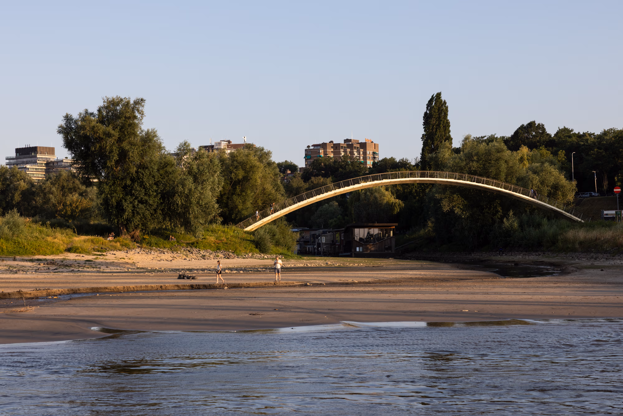 Mensen lopen over een strandje aan de Waal in Nijmegen waar normaal water staat. Aan de brug en de woonboten op de achtergrond is te zien tot waar het water normaal gesproken komt.