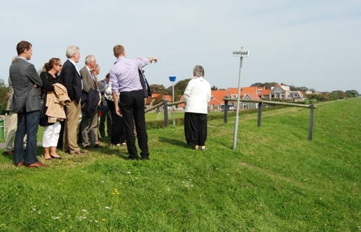 De deltacommissaris (3e van links) luistert op de dijk naar een toelichting door de burgemeester van Vlieland, Yorick Haan.