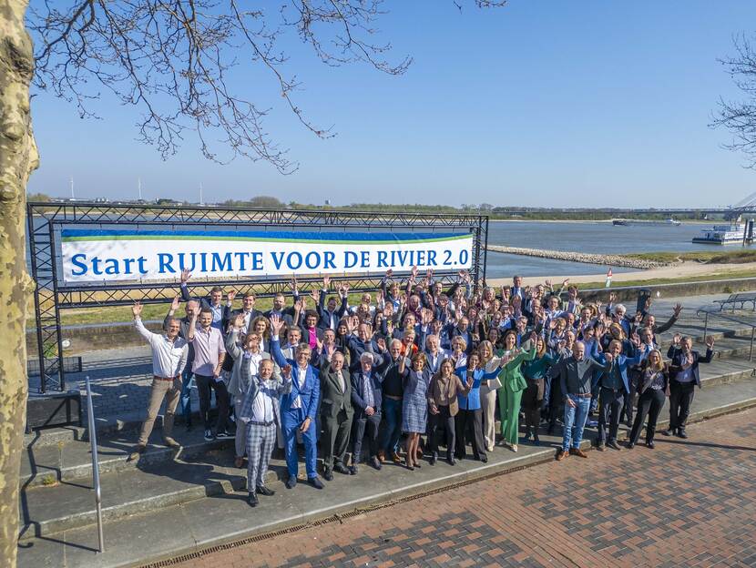 Alle samenwerkende partijen staan voor de banner met tekst “Start RUIMTE VOOR DE RIVIER 2.0” en zwaaien lachend naar de camera.